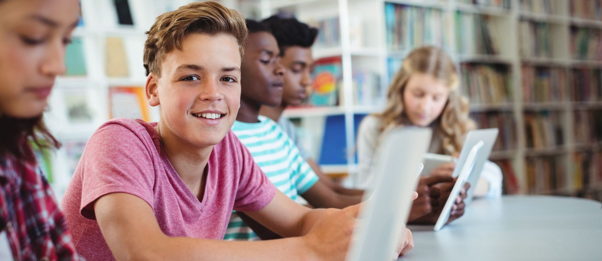 A group of young people are sitting at a table in a library using laptops and tablets.