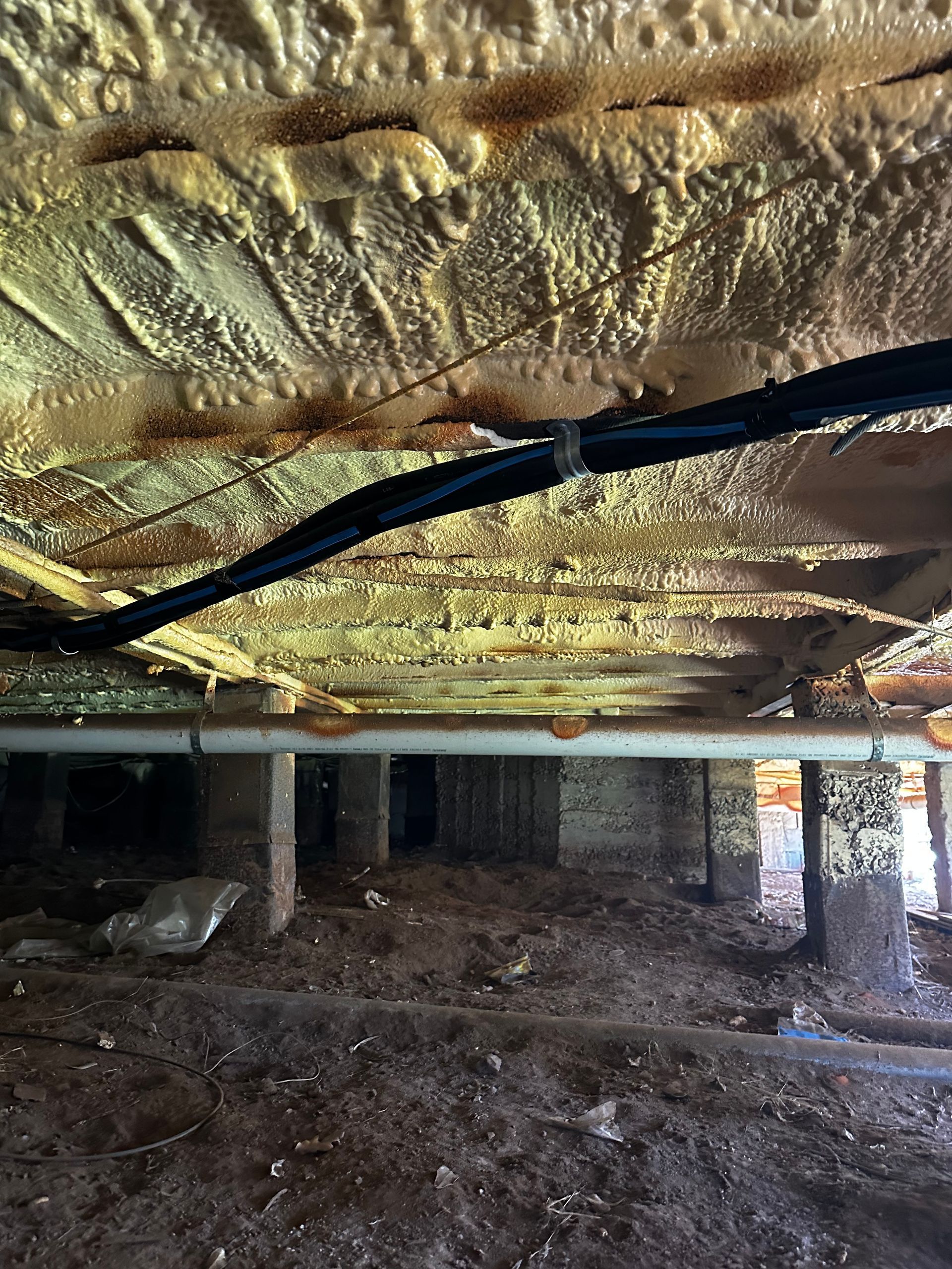 Underside of a house foundation with spray foam insulation, pipes, wiring, and dirt floor  — Proshield Insulation & Waterproofing In Shell Cove, NSW