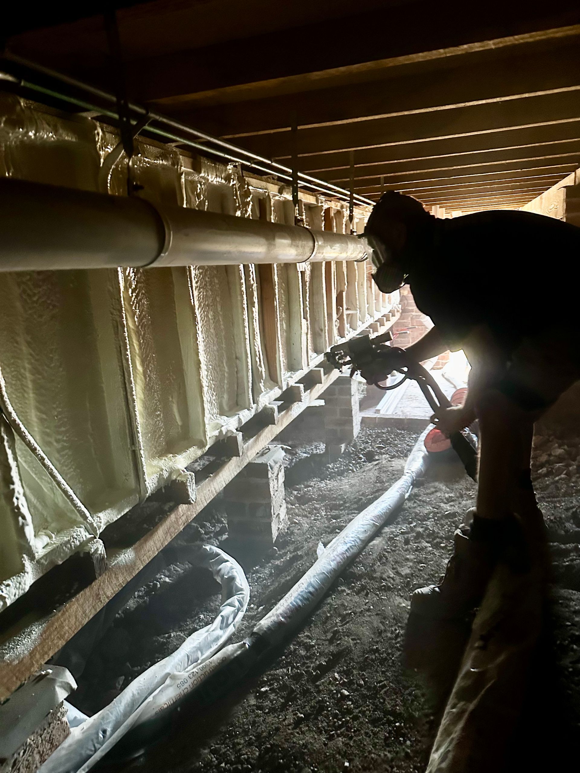 Person spraying foam insulation in a crawl space. Beige foam covers walls, pipes, and beams  — Proshield Insulation & Waterproofing In Nowra, NSW