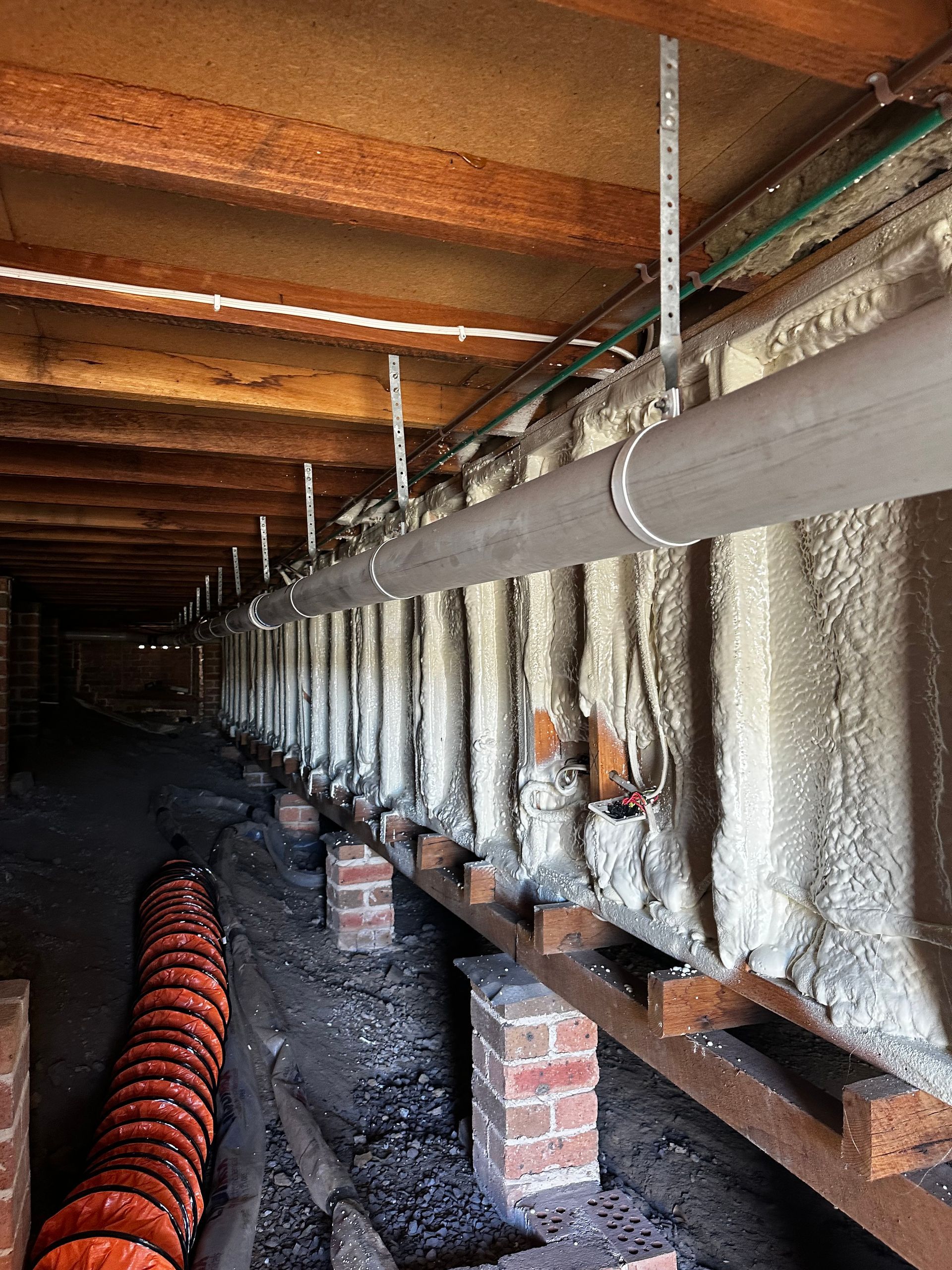 Underneath a house, a long grey pipe is supported by vertical metal rods. Insulation covers the side walls  — Proshield Insulation & Waterproofing In Shell Cove, NSW