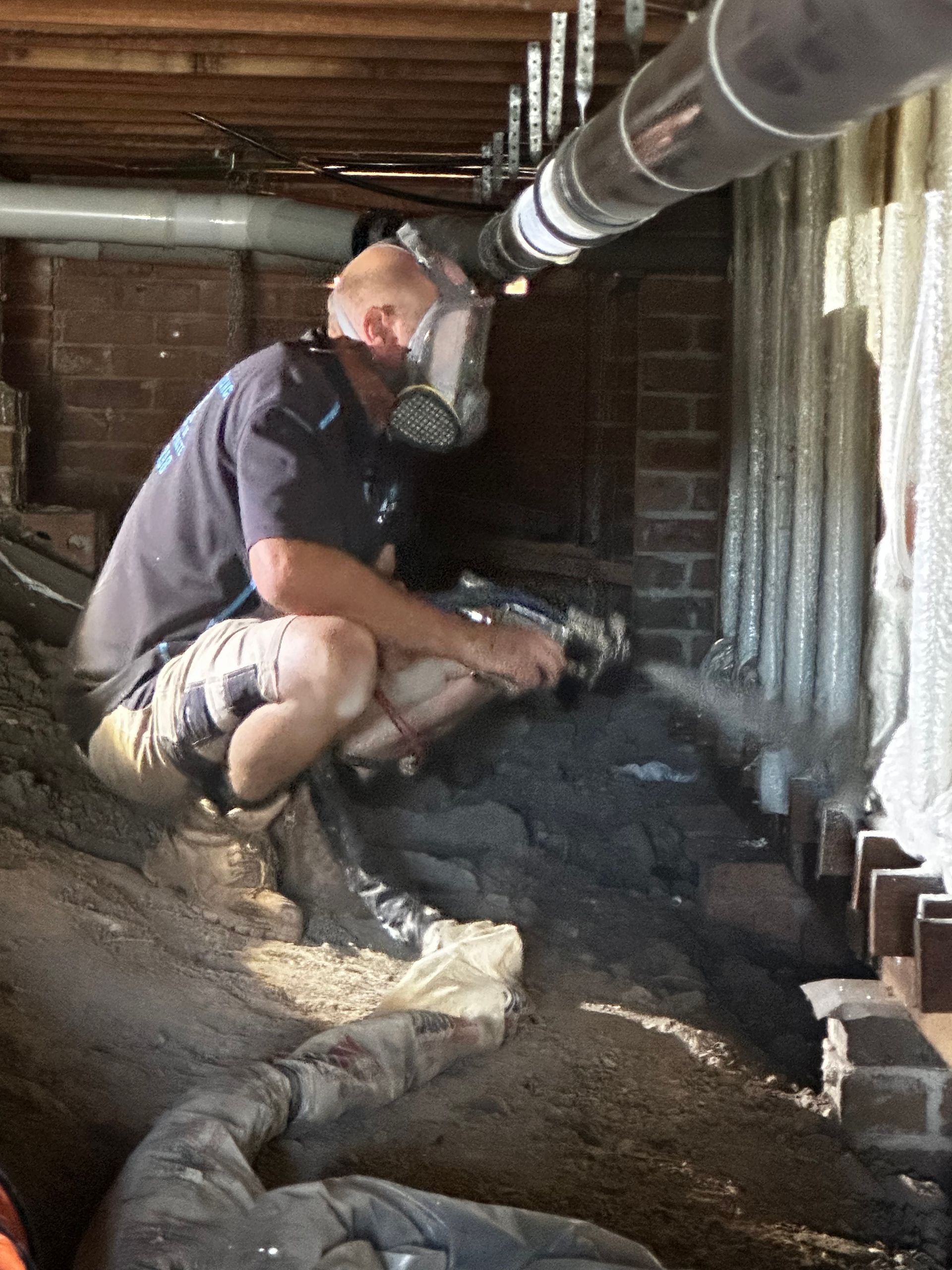 Man in respirator kneels in crawl space, working with gray material; ventilation tube overhead  — Proshield Insulation & Waterproofing In Shell Cove, NSW