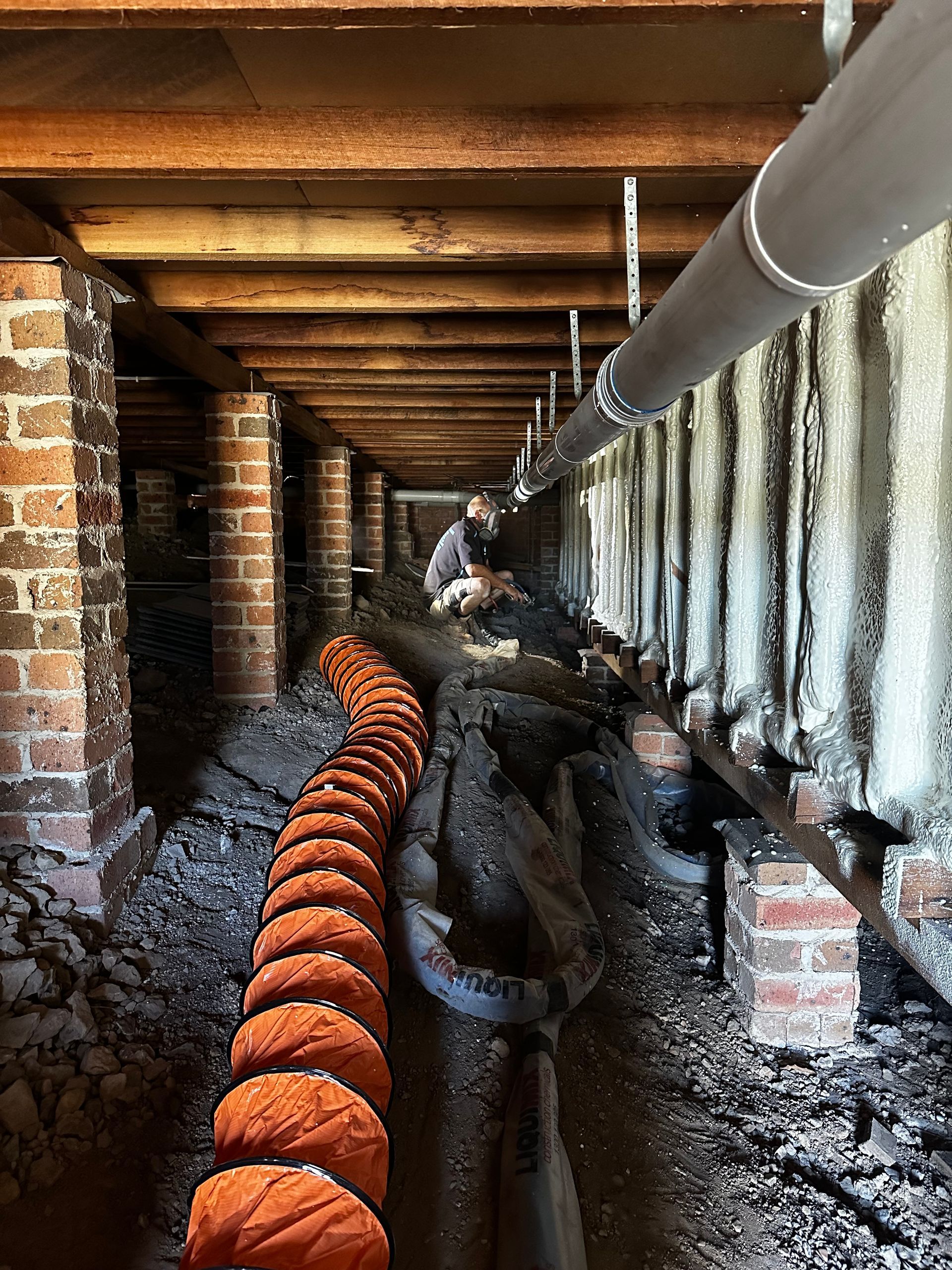 Man working in crawl space with brick pillars, pipes, and orange insulation — Proshield Insulation & Waterproofing In Campbelltown, NSW