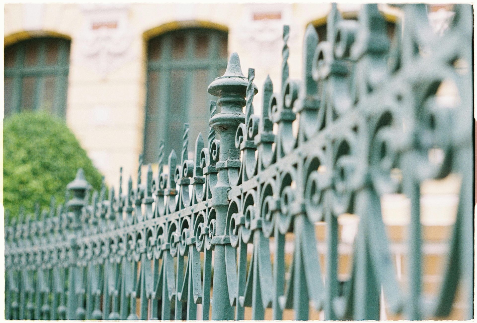 A close-up, angled view of a decorative green wrought iron fence in front of a blurred historical building.