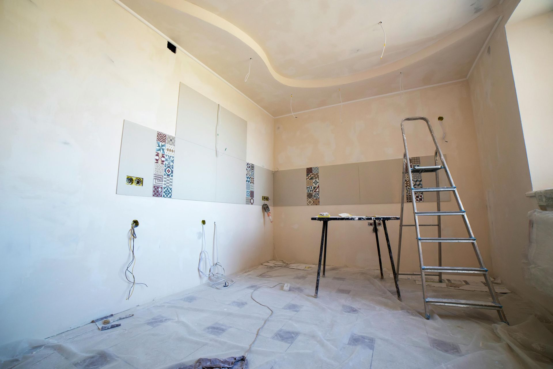 An unfinished kitchen renovation with a metal stepladder, a small table, and partially tiled walls in a bright room.