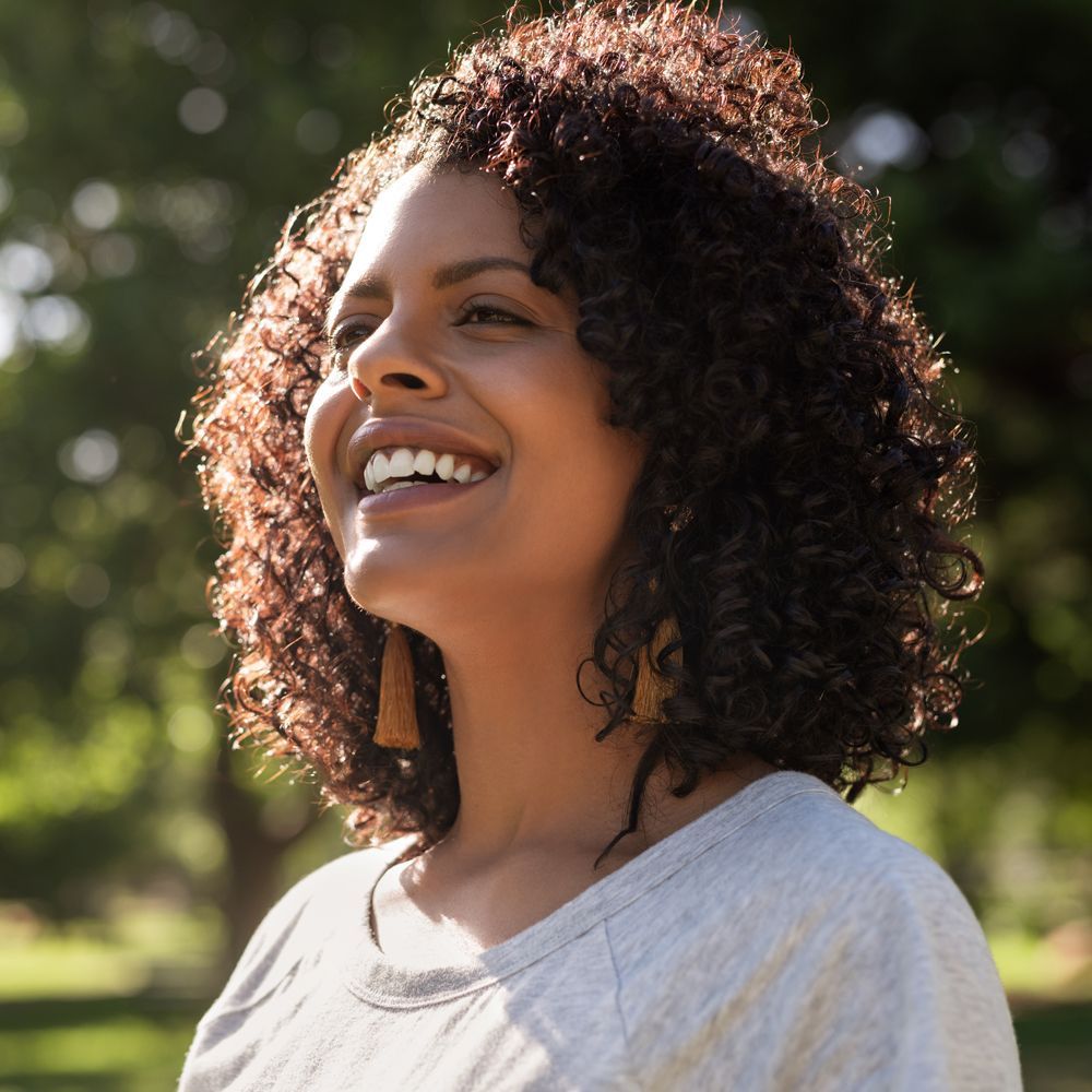 A woman with curly hair is smiling in a park.