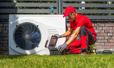 Technician in red uniform kneels beside outdoor HVAC unit, inspecting the control panel in a yard.