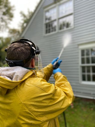 Person in yellow rain jacket pressure-washing a gray house exterior.