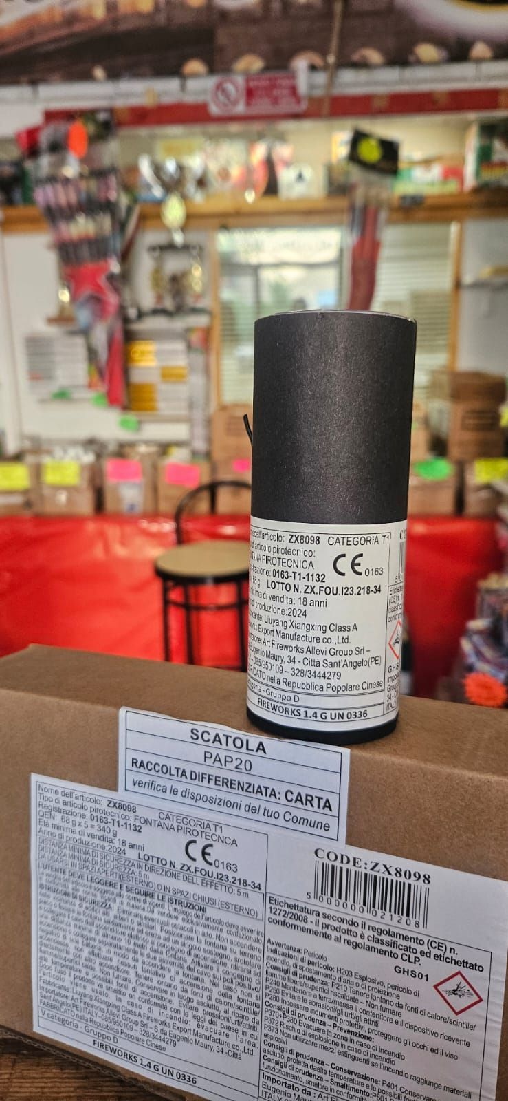 Un oggetto cilindrico nero con un'etichetta è appoggiato su una scatola di cartone. Sullo sfondo, un negozio al dettaglio.