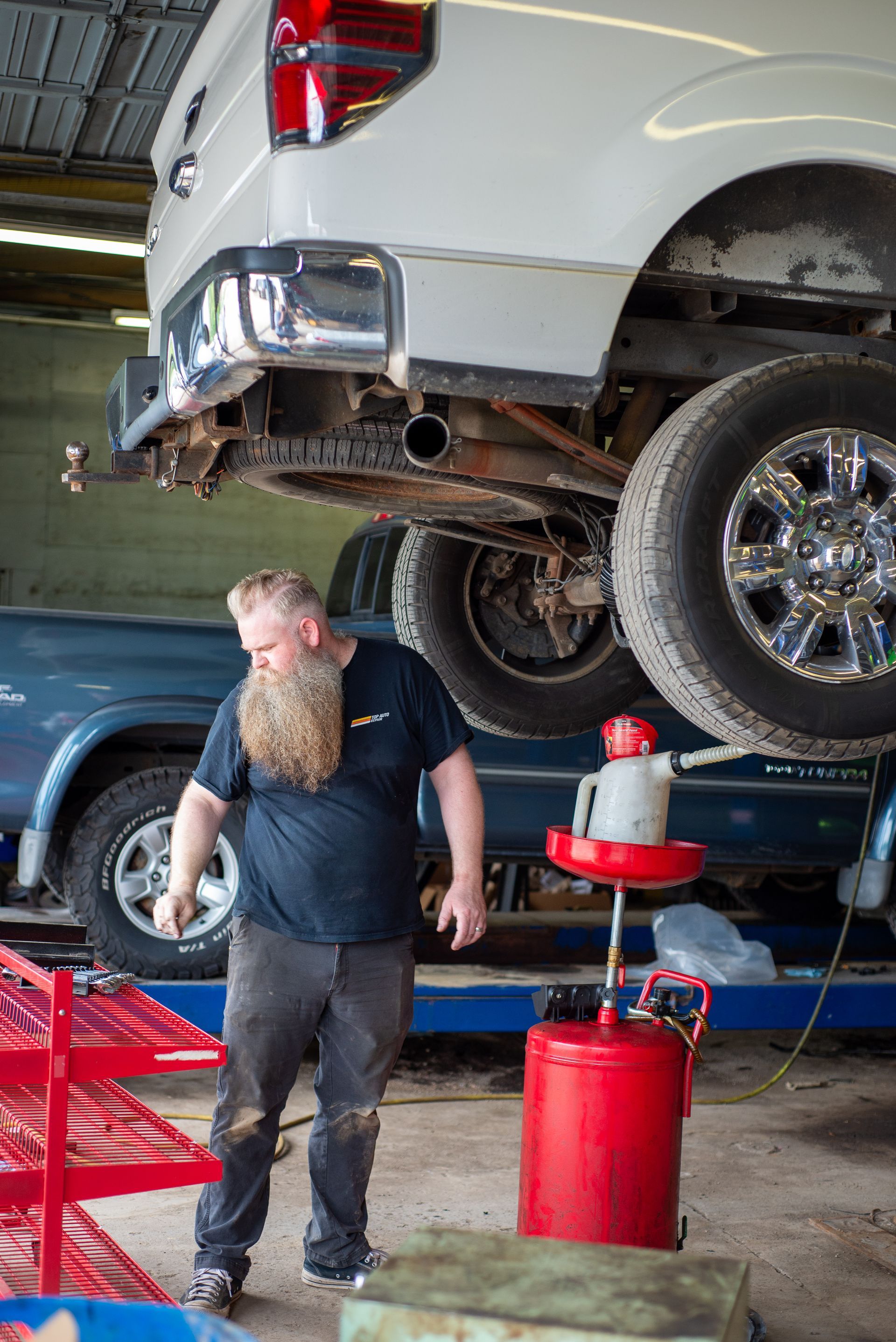 A man with a beard is standing under a truck in a garage.