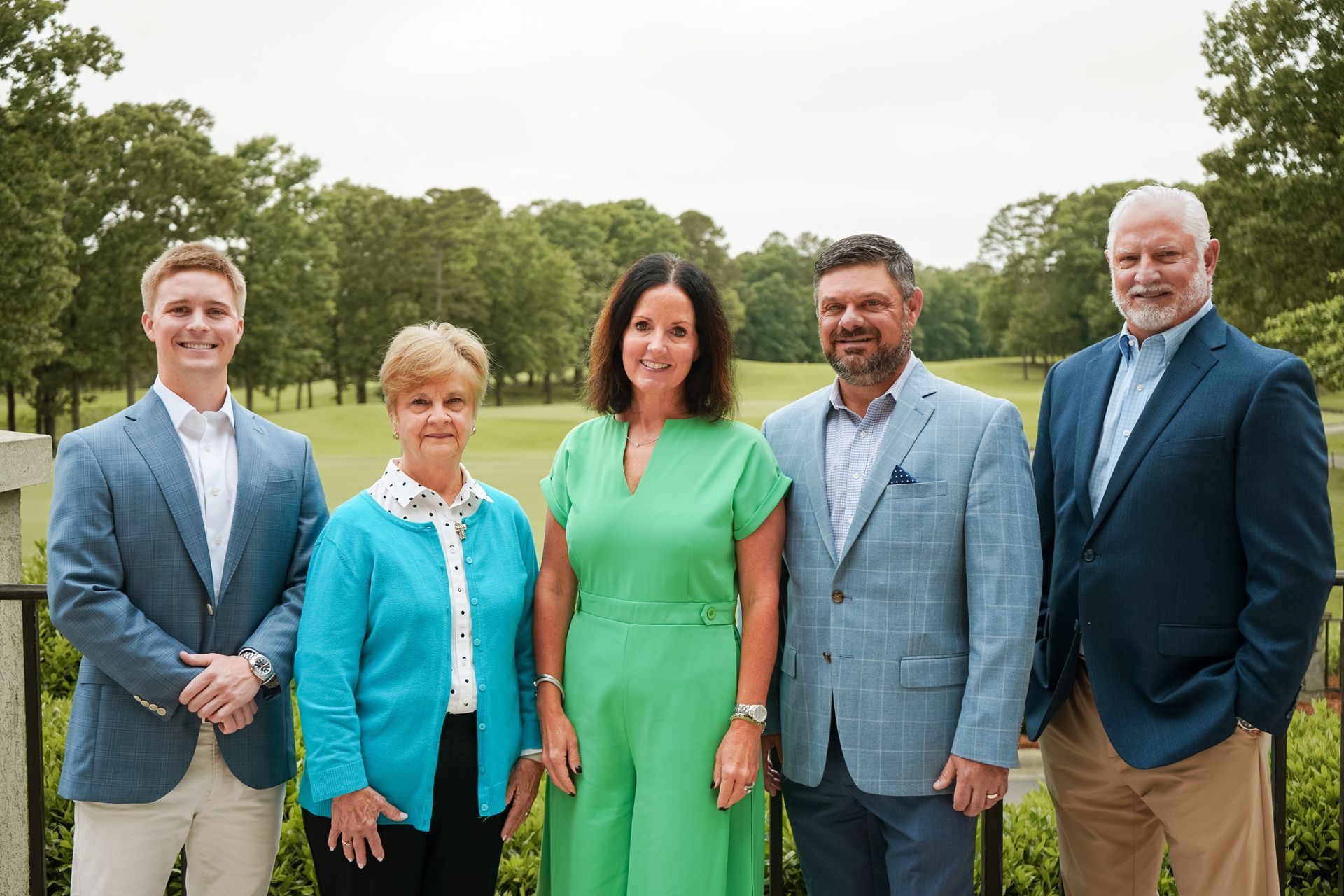 Five people standing outdoors in a park, dressed in formal and business attire.