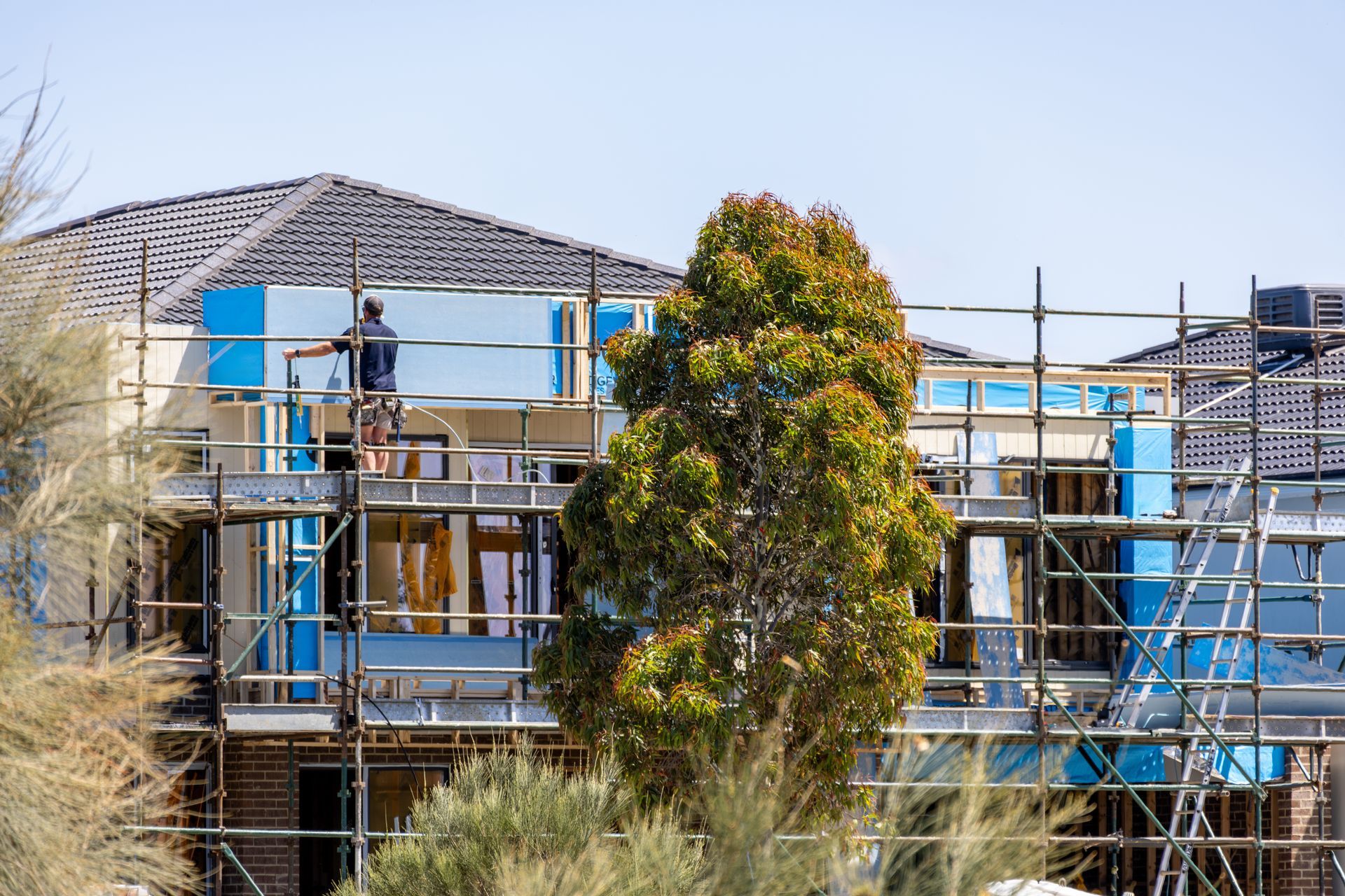 Construction workers on scaffolding beside a building under renovation, with blue tarps and trees in front