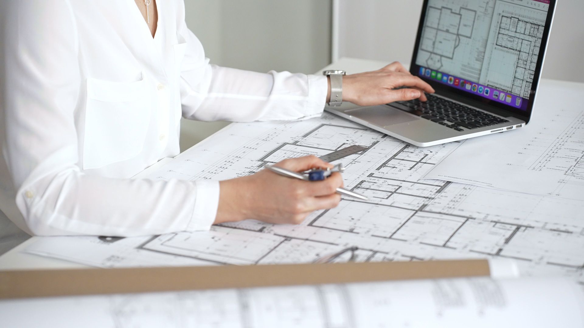 Person reviewing architectural blueprints on a desk while using a laptop with floor plans on screen