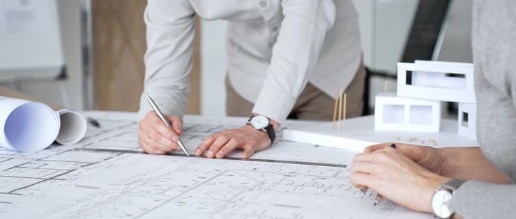 Hands drawing architectural plans on a desk with a white scale model in a bright office
