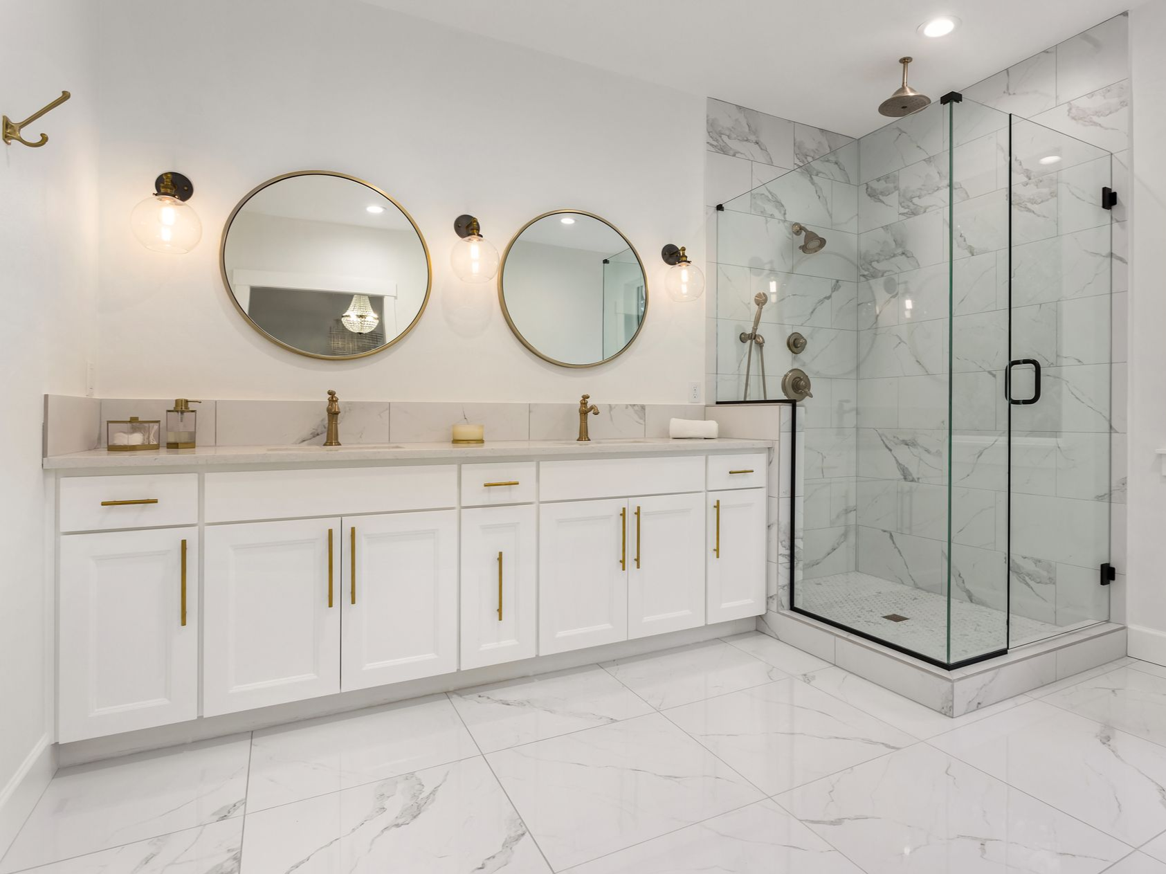 Modern white bathroom with double vanity, gold fixtures, circular mirrors, and a glass-enclosed marble shower.