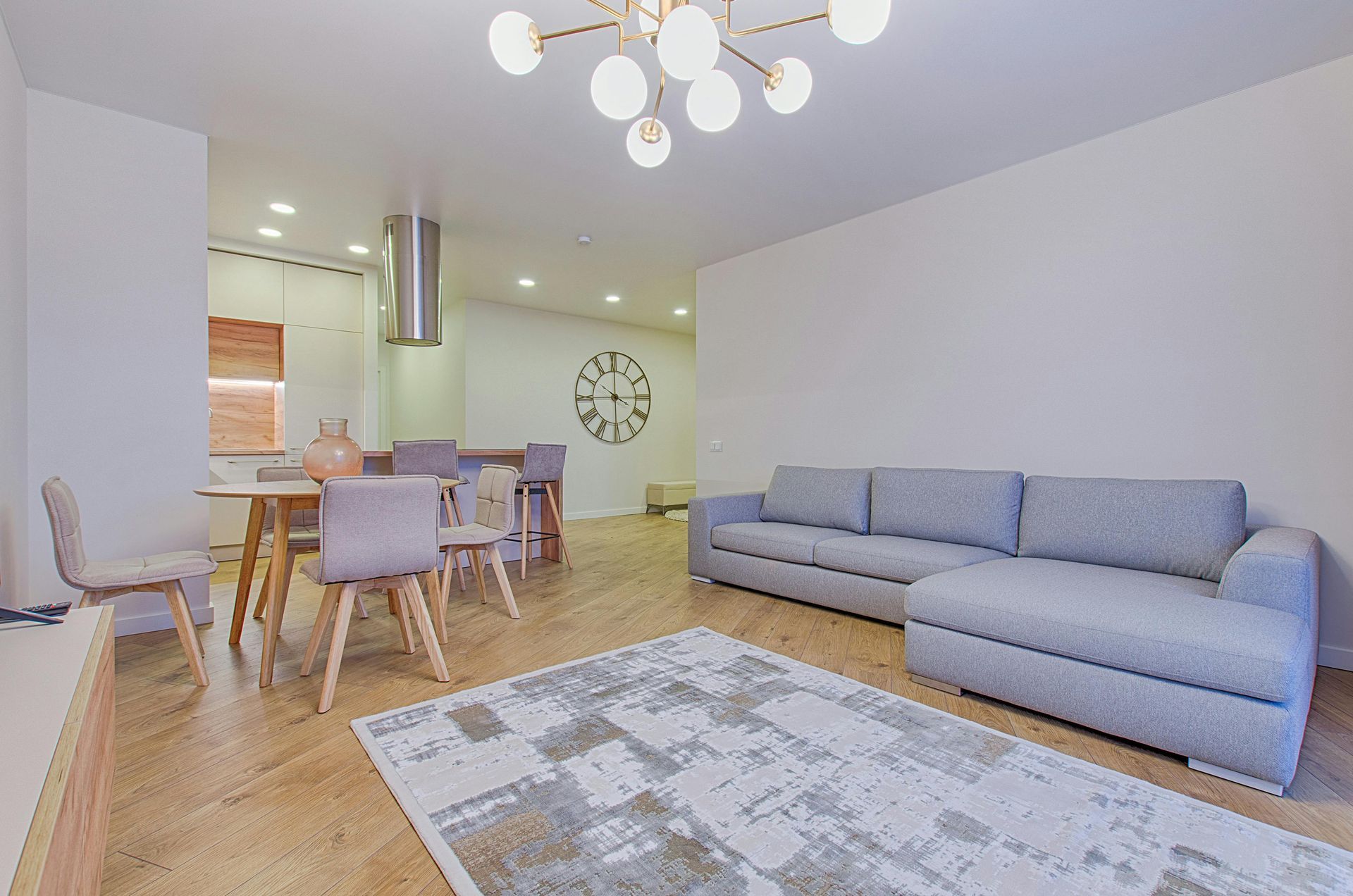 Living room with a light gray sectional sofa, dining table, and rug on a hardwood floor, with a modern chandelier.