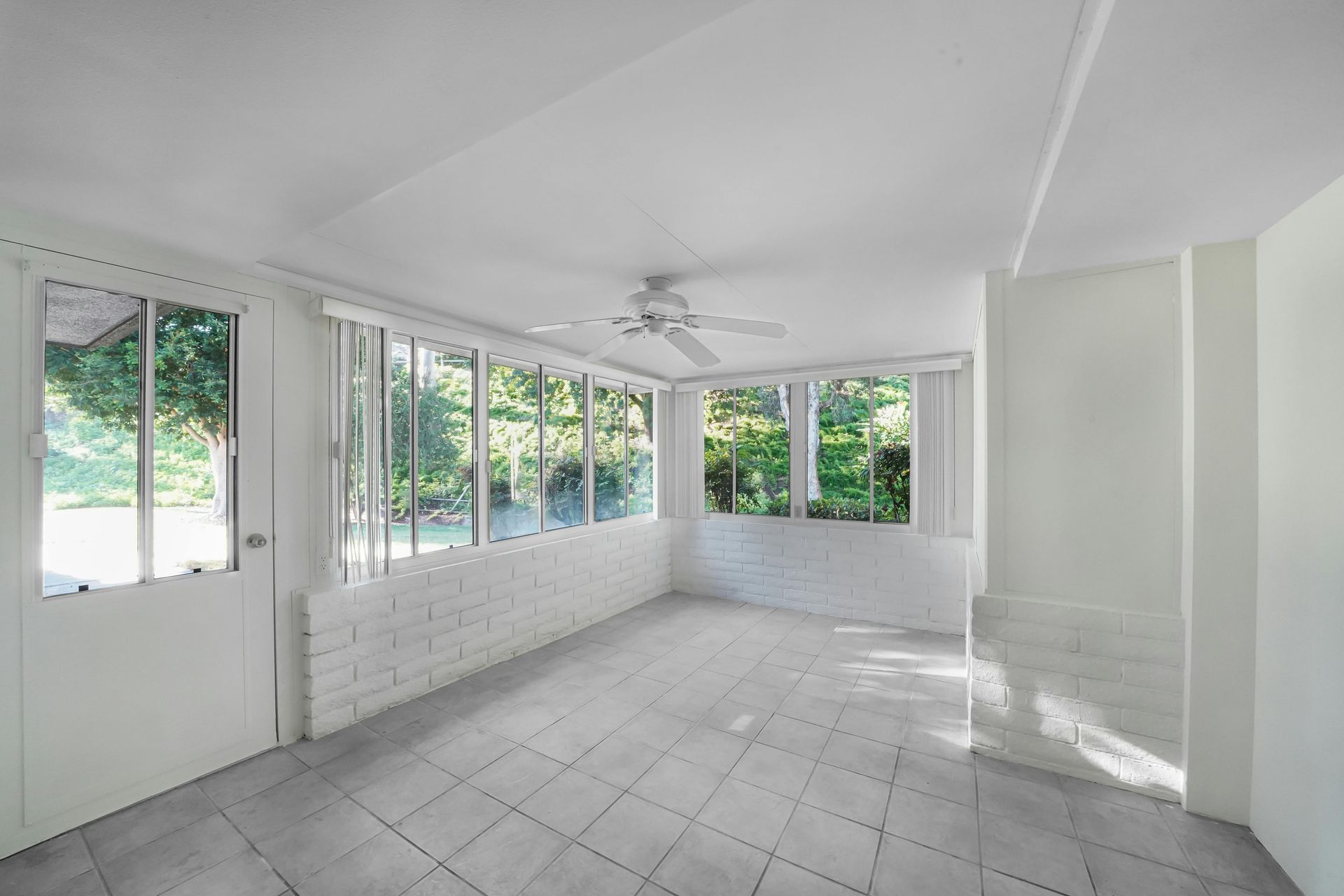Bright, empty sunroom with white brick, large windows, and a ceiling fan.