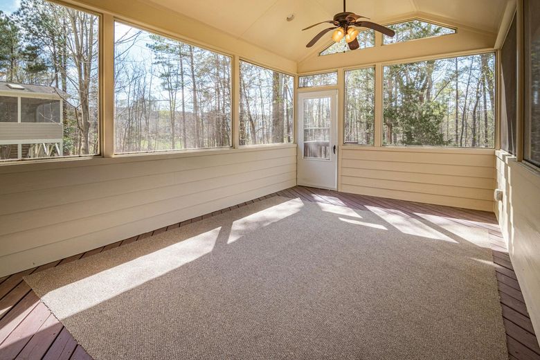 Screened porch with light tan walls, carpet, and a ceiling fan, with a door and windows overlooking trees.