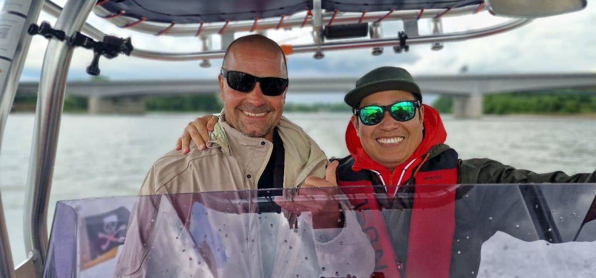 Zwei Männer stehen nebeneinander auf einem Boot und halten Fische in den Händen.