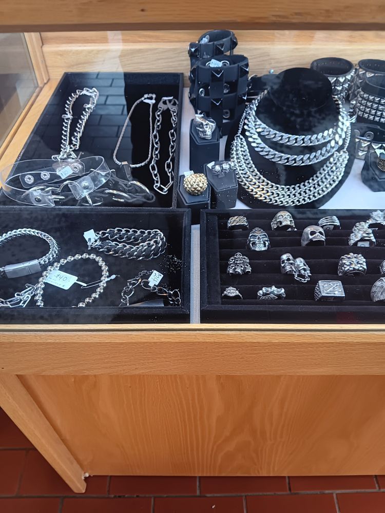 A display case containing various silver-toned chains, rings, and spiked leather cuffs on velvet-lined trays.