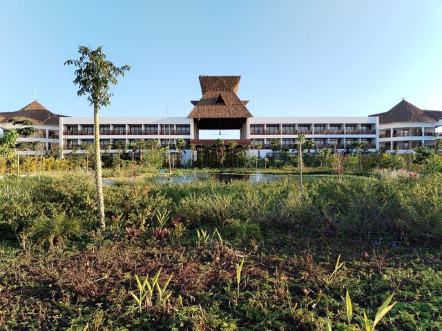 Amplio edificio de un complejo turístico con techo central de paja, rodeado de vegetación bajo un cielo azul despejado.