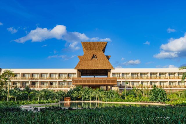 Amplio edificio estilo resort con una torre central de techo de paja, ubicado en medio de una exuberante vegetación bajo un cielo azul brillante.