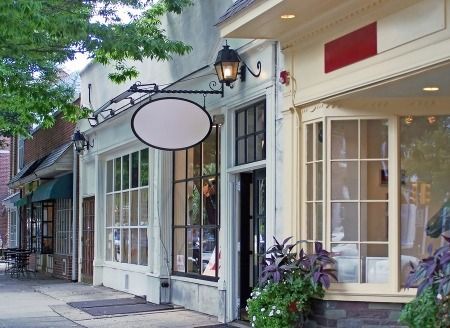 Shops on a sunny street with a blank oval sign, glass windows, and plants in front.