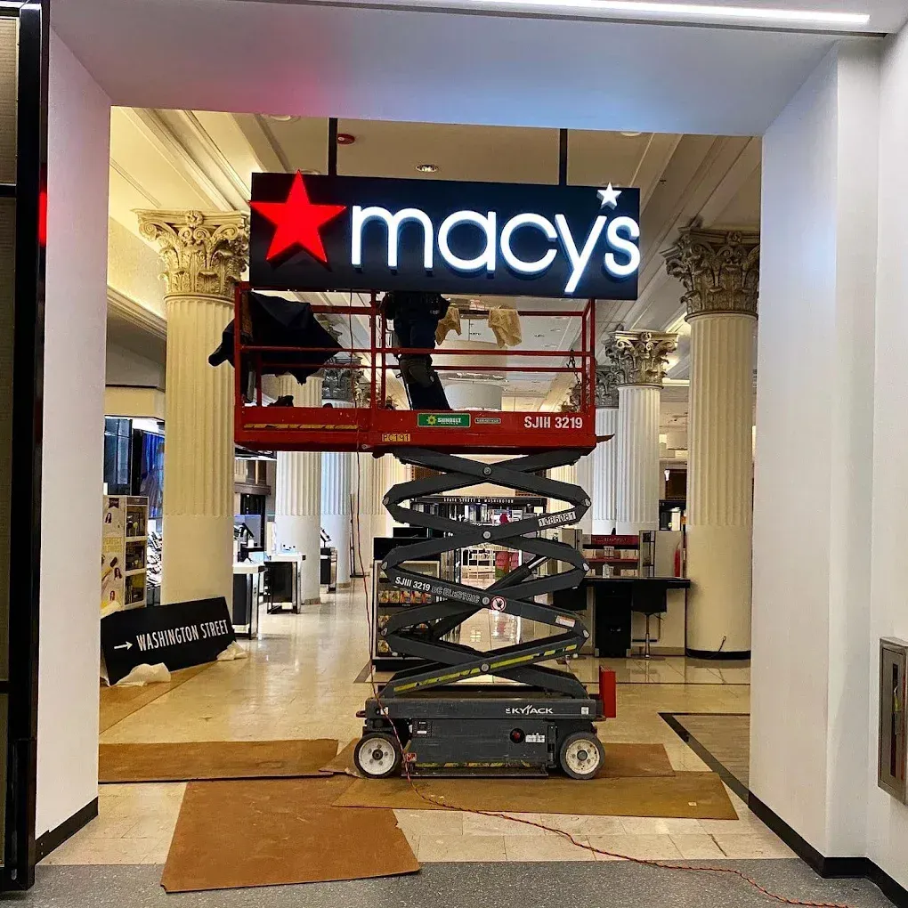 A worker on a scissor lift installs a Macy's sign indoors, with a red star and white letters.