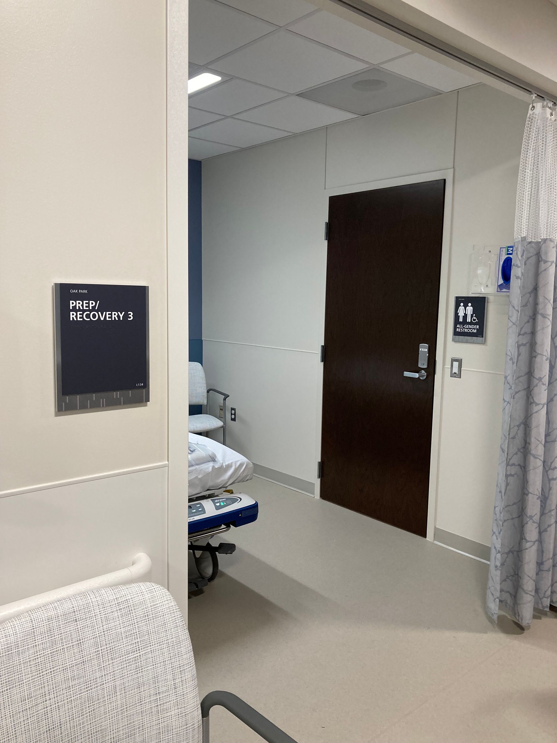 Hospital hallway, entrance to room with sign, a gurney, seating, and restroom.