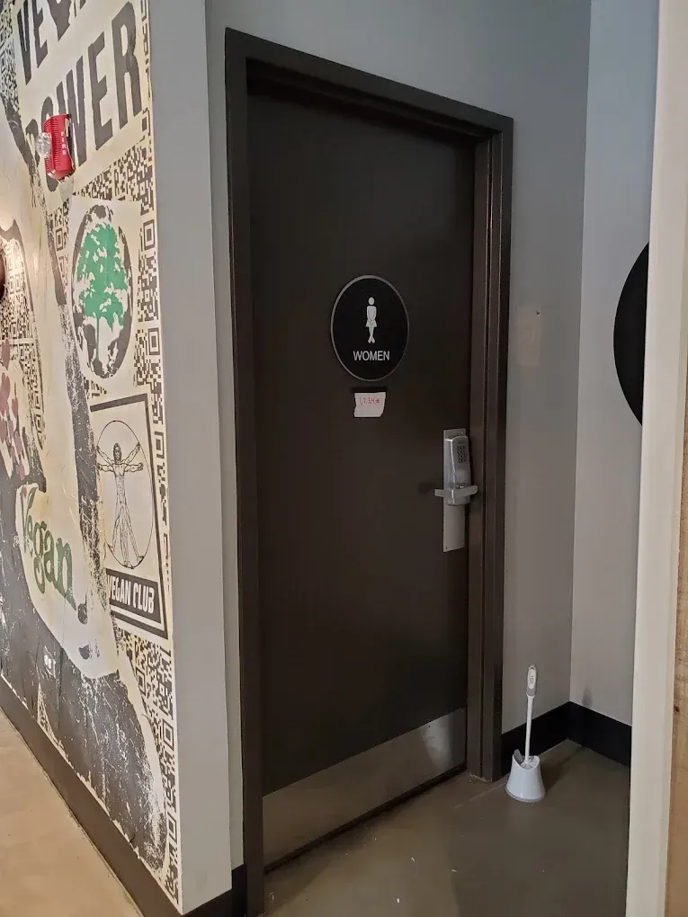 Women's restroom door with a black silhouette symbol, gray door, and poster-covered wall to the left.