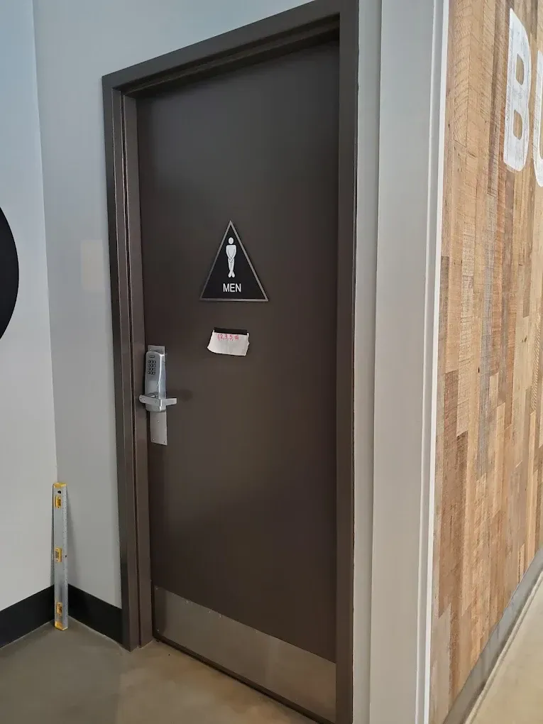 Dark gray restroom door with triangular gender symbol, handle, and note. Adjacent to a wood-paneled wall.