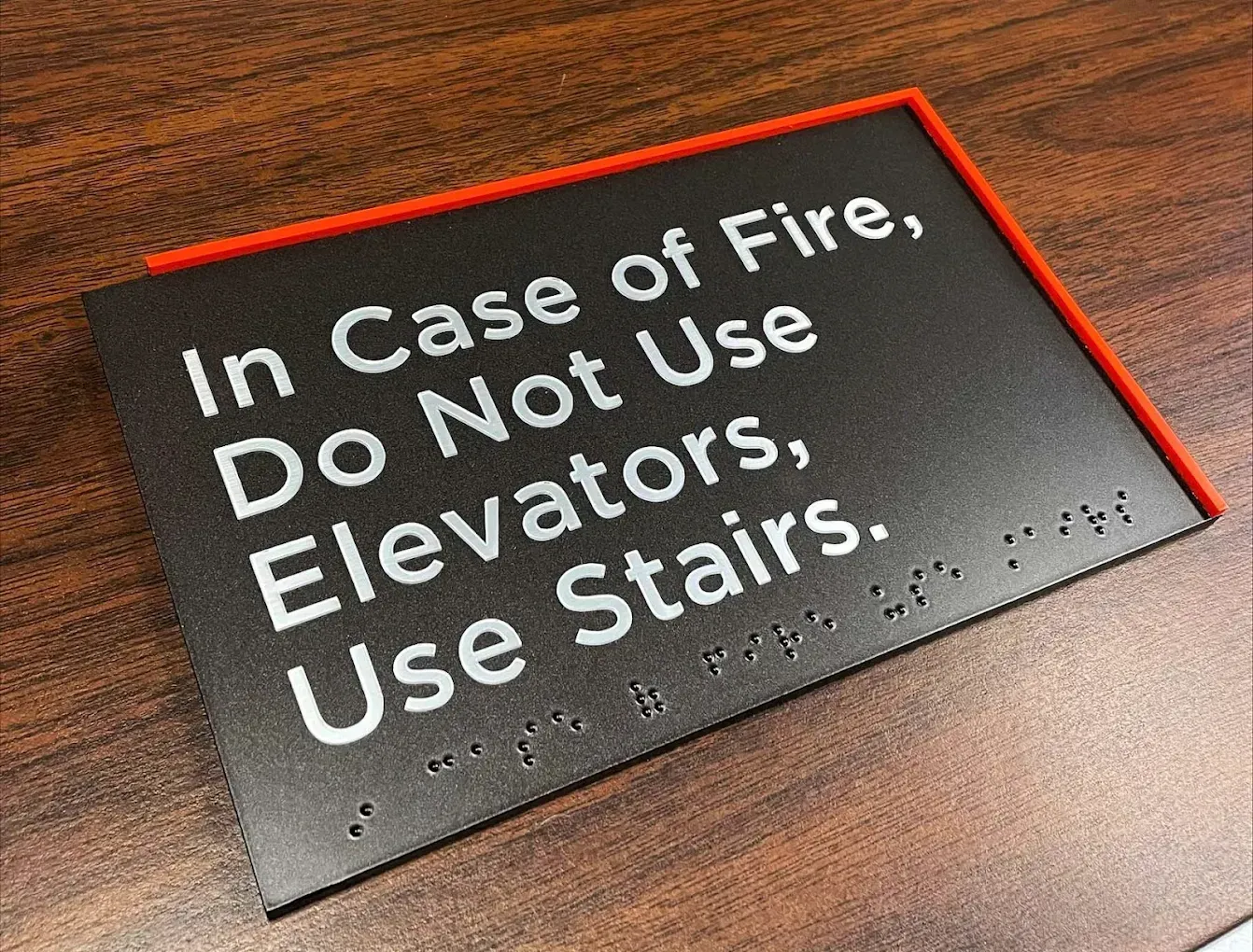 Sign: "In Case of Fire, Do Not Use Elevators, Use Stairs." Includes Braille, red border, on a wooden surface.