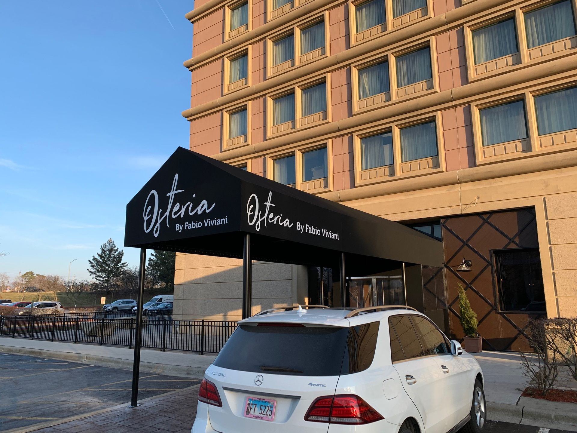 White SUV parked in front of an Ostera restaurant entrance, under a black awning. Building is beige with square windows.
