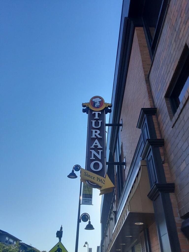 Sign for "Turano" restaurant with an arrow pointing toward the entrance, against a blue sky.