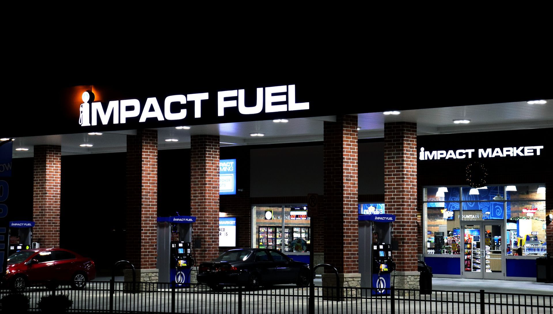 Night shot of an Impact Fuel gas station with illuminated sign, gas pumps, and cars filling up.