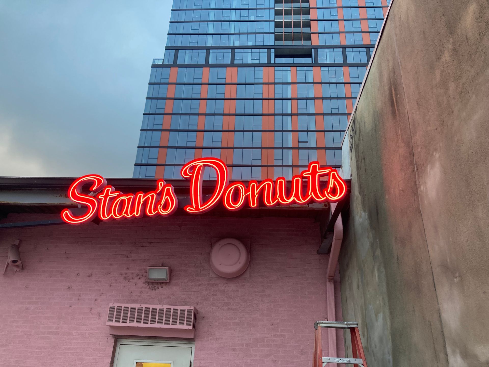 Neon sign for Stan's Donuts on a pink building, with a tall modern building in the background.