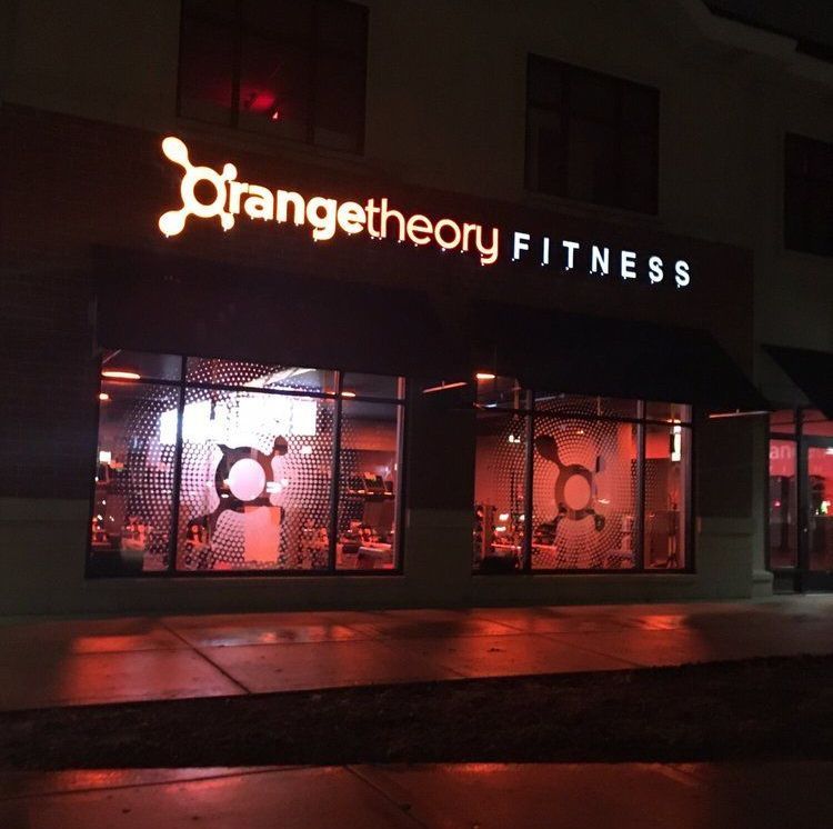 OrangeTheory Fitness storefront at night, illuminated in orange, logo above windows.