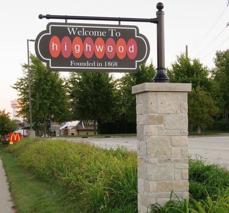 Sign welcoming visitors to Highwood, founded in 1868, mounted on a stone post with McDonald's visible in the background.
