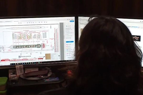 Person with dark hair looking at two computer monitors displaying schematics, indoors.