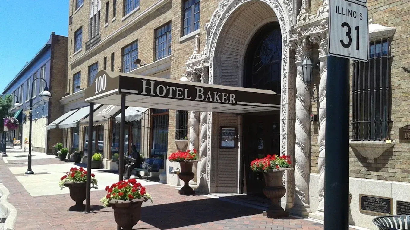 Hotel Baker, St. Charles, Illinois, entrance with awning. Beige building, red flowers, blue sky, and a street sign.