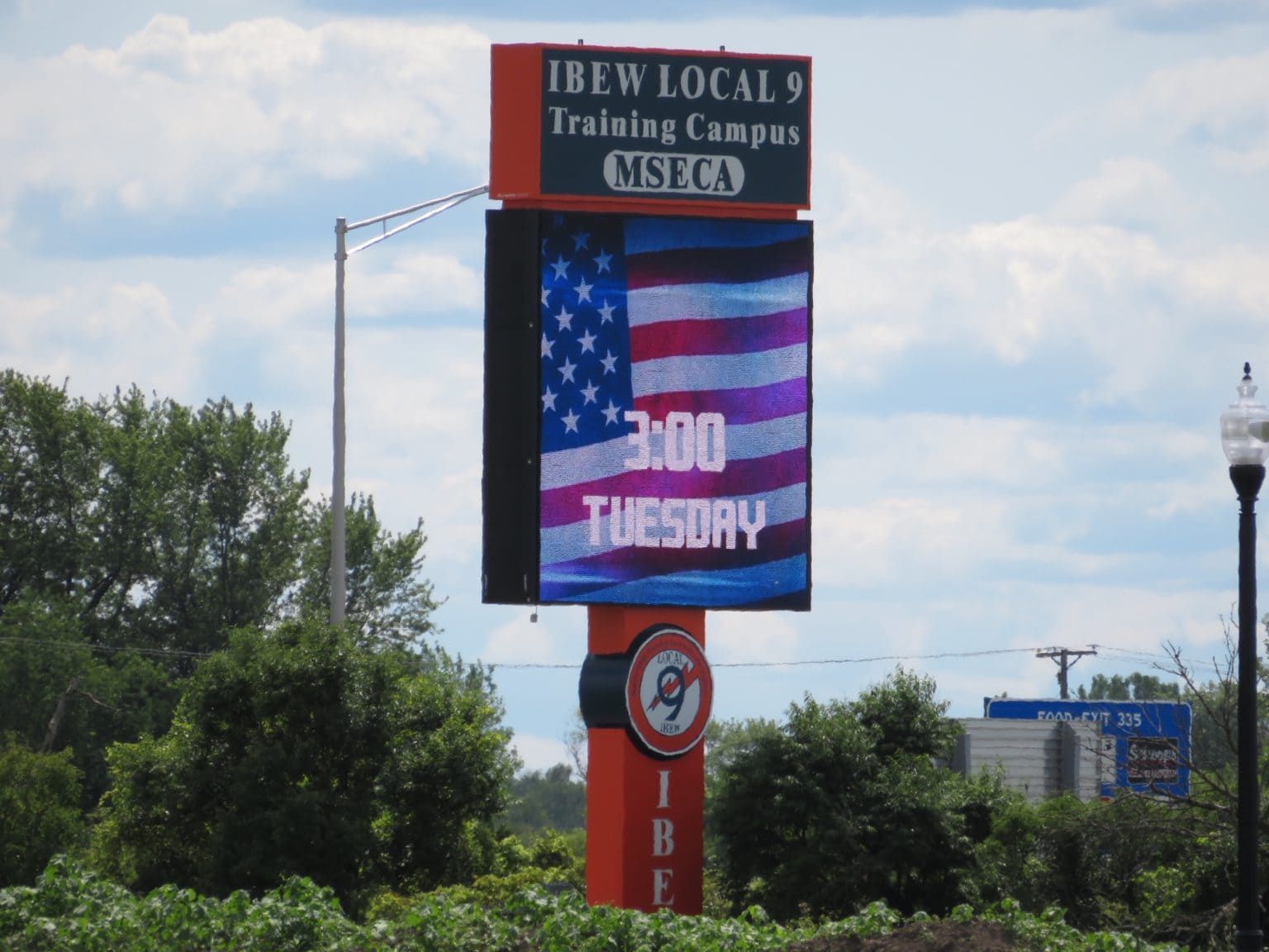 Sign for IBEW Local 9 training campus showing an American flag, time, and day against a cloudy sky.