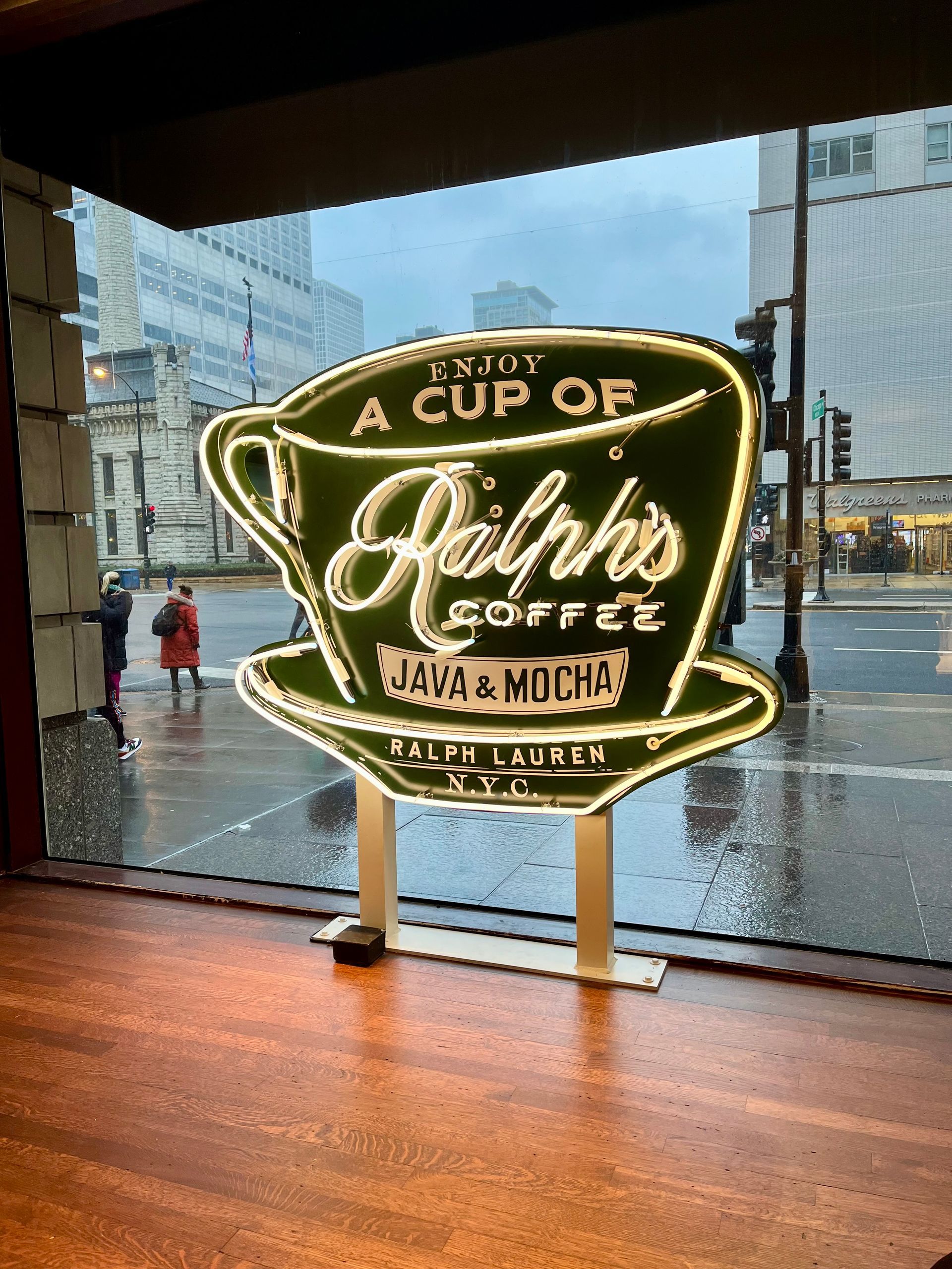 Green neon sign for Ralph's Coffee, shaped like a coffee cup, inside a store window. Rainy street visible outside.