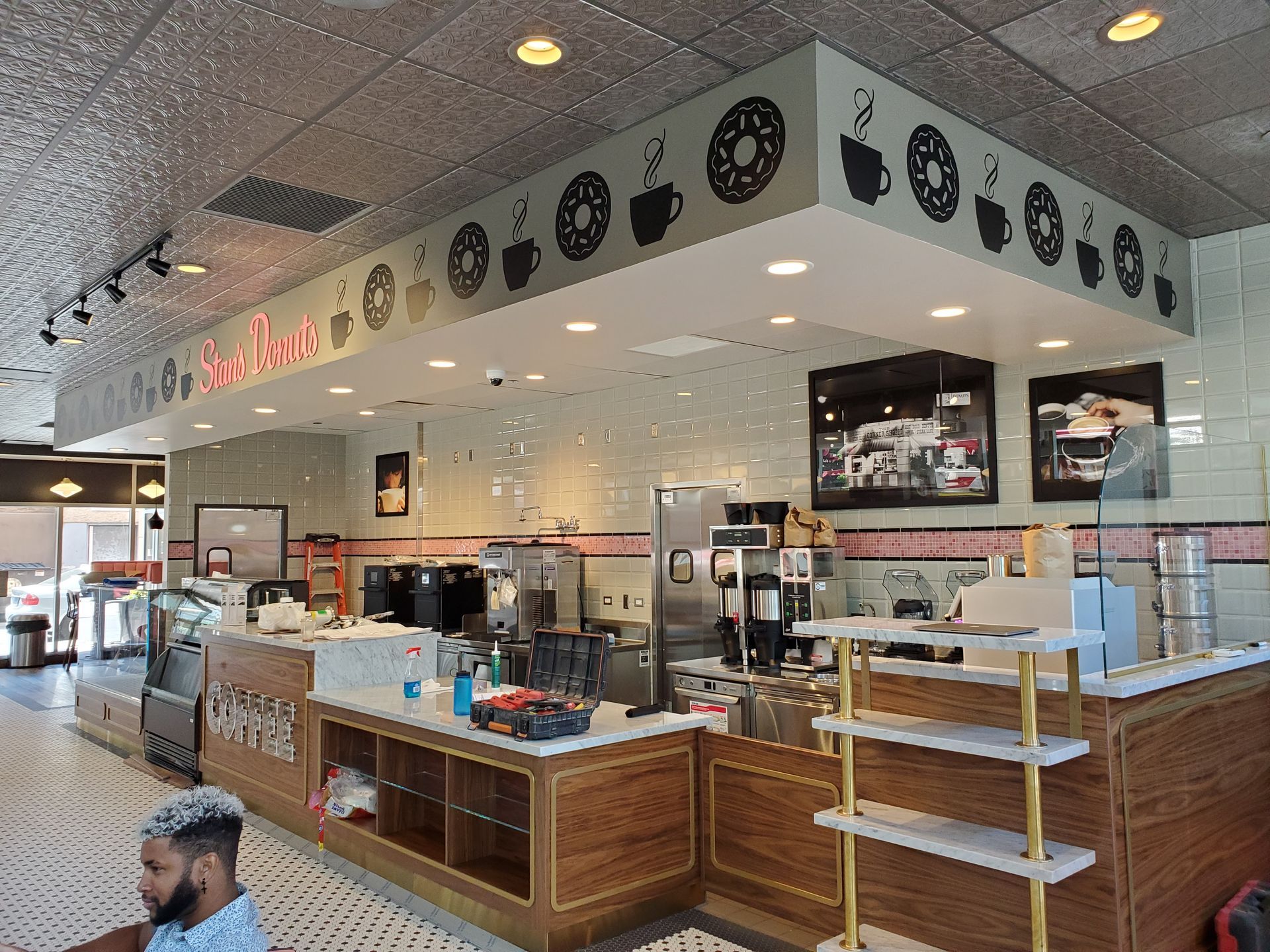 Interior of a donut shop: counter with donuts and coffee, decorative ceiling, tile walls, and a customer.