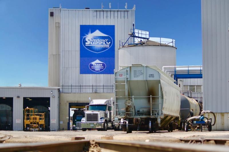 Truck and train car loading at a factory with large blue logo on a bright, sunny day.