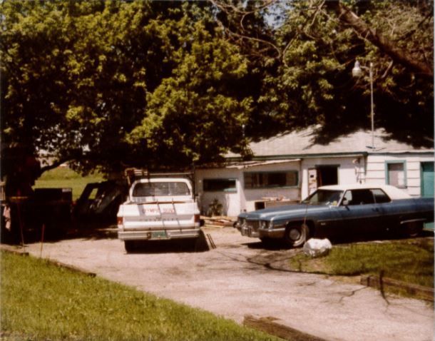 A white pickup truck and blue car parked in front of a white house with a large tree.
