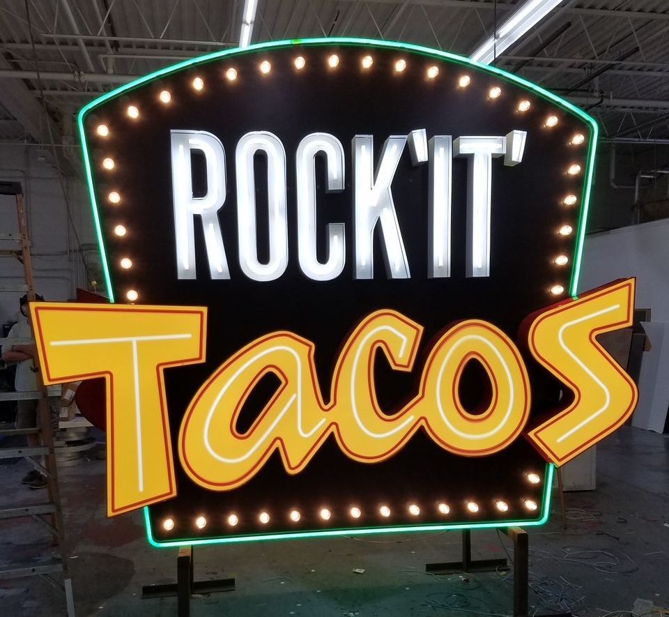 Rock'it' Tacos sign with neon lights: black background, white text, yellow tacos, green and yellow border, indoors.