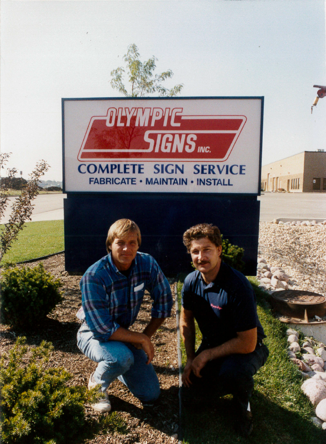 Two men kneeling in front of a sign for Olympic Signs, a sign company.