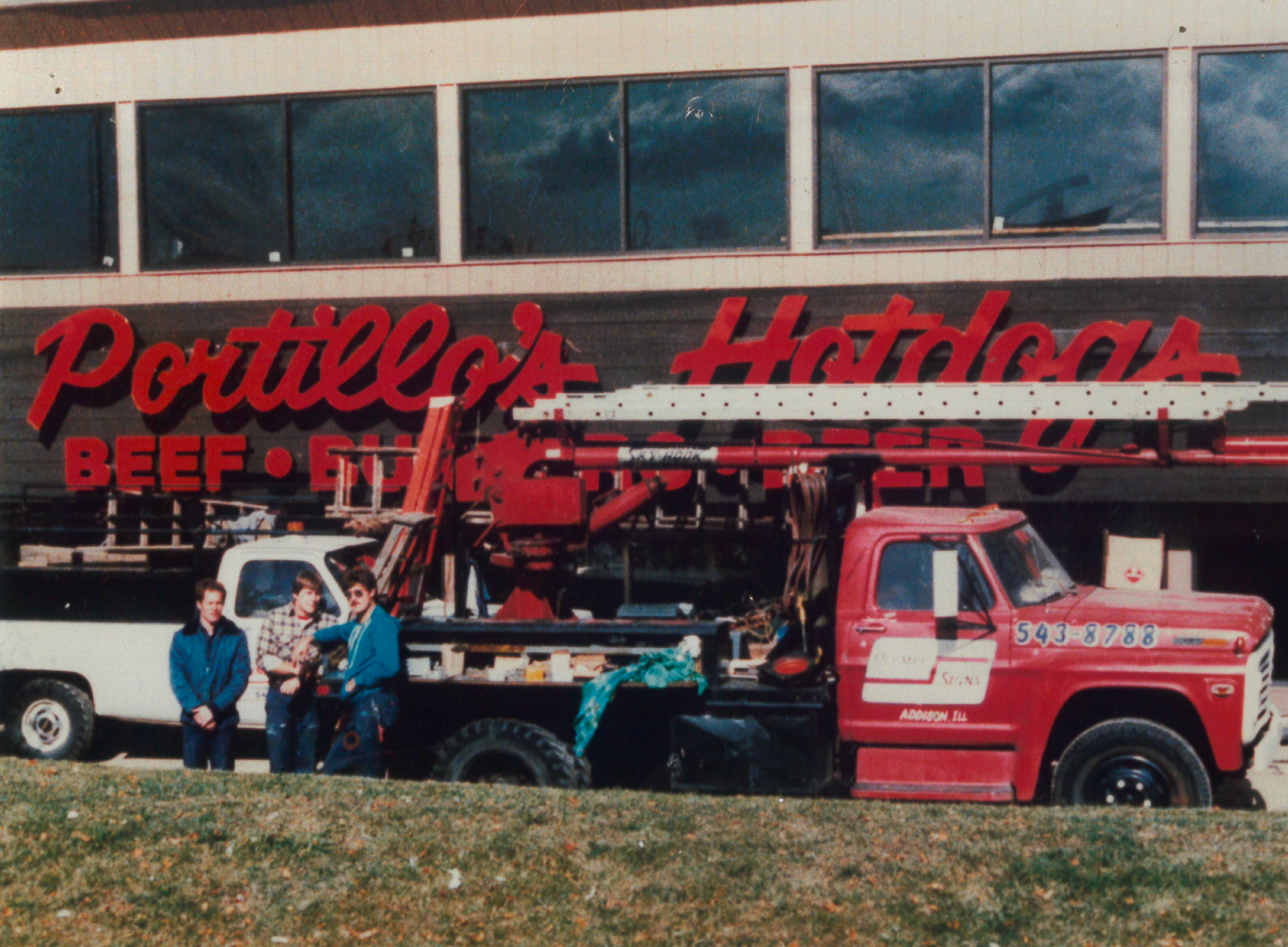 Men standing near a red truck with a ladder lift in front of a Portillo's Hot Dogs restaurant sign.
