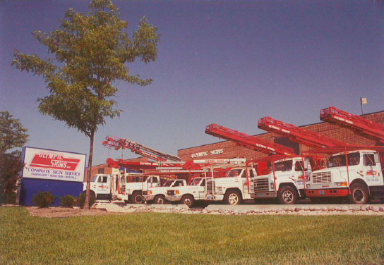 White and red service trucks parked outside a business with a sign on a sunny day.