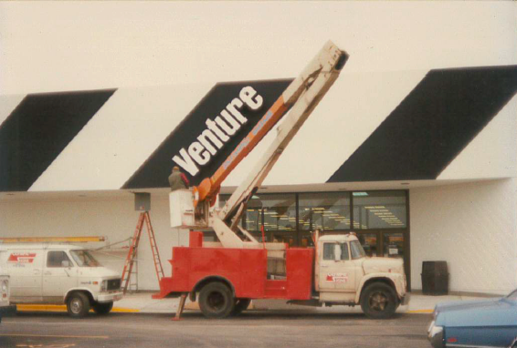A red and white boom truck outside a Venture store, working on the building's signage.