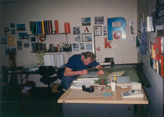 Man leans over a drafting table in a cluttered office, working on a project. Letter art on wall.